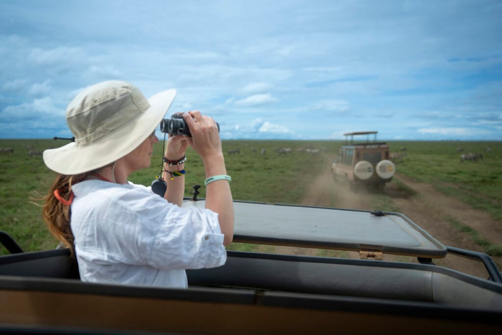 Woman with hat and binoculars standing in a jeep on a road in Tanzania